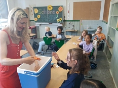 Auf dem Bild sieht man einen Klassenraum. In diesem verteilt eine Frau links im Bild Eis an Schülerinnen und Schüler.