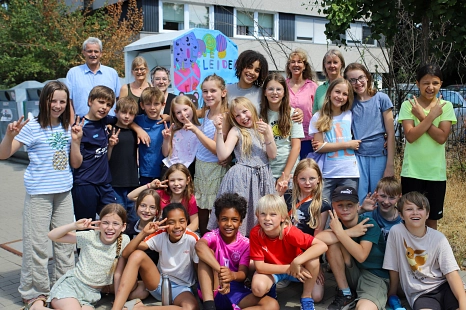 In dem Bild sieht man einige Kinder, die vor einem bemalten Altkleider-Container stehen.