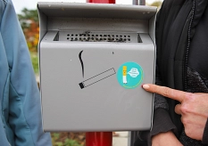 In Nahaufnahme der neue Aschenbecher an der Bushaltestelle im Waldtal. Er ist grau und hat ein Zigarettensymbol vorne drauf.