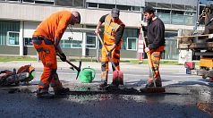 Drei Männer in orangefarbener Kleidung füllen mit Hacke und Schippe ein Schlagloch in der Straße auf.