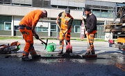 Drei Männer in orangefarbener Kleidung füllen mit Hacke und Schippe ein Schlagloch in der Straße auf.