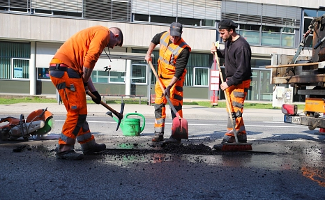Drei Männer in orangefarbener Kleidung füllen mit Hacke und Schippe ein Schlagloch in der Straße auf.