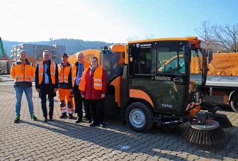Fünf Menschen stehen vor der neuen elektrischen Kehrmaschine. Sie blicken in die Kamera und tragen orangene Warnwesten.