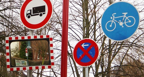 Das Foto zeigt verschiedene Verkehrsschilder und einen Spiegel an einem Mast.
